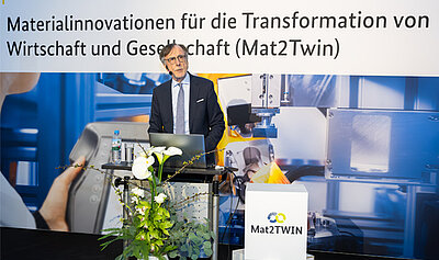 Ein Mann steht an einem Podium auf der Bühne. Auf dem Banner im Hintergrund steht "Materialinnovation für die Transformation von Wirtschaft und Gesellschaft"