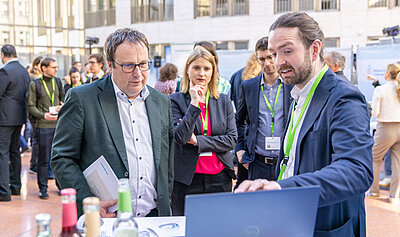 Mehrere Personen stehen bei der Ausstellung um einen Laptop. Ein Mann zeigt darauf und erklärt etwas.