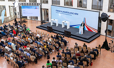 Blick von oben auf das Publikum und die Bühne. Am Rednerpult auf der Bühne steht NRW-Umweltminister Oliver Krischer.